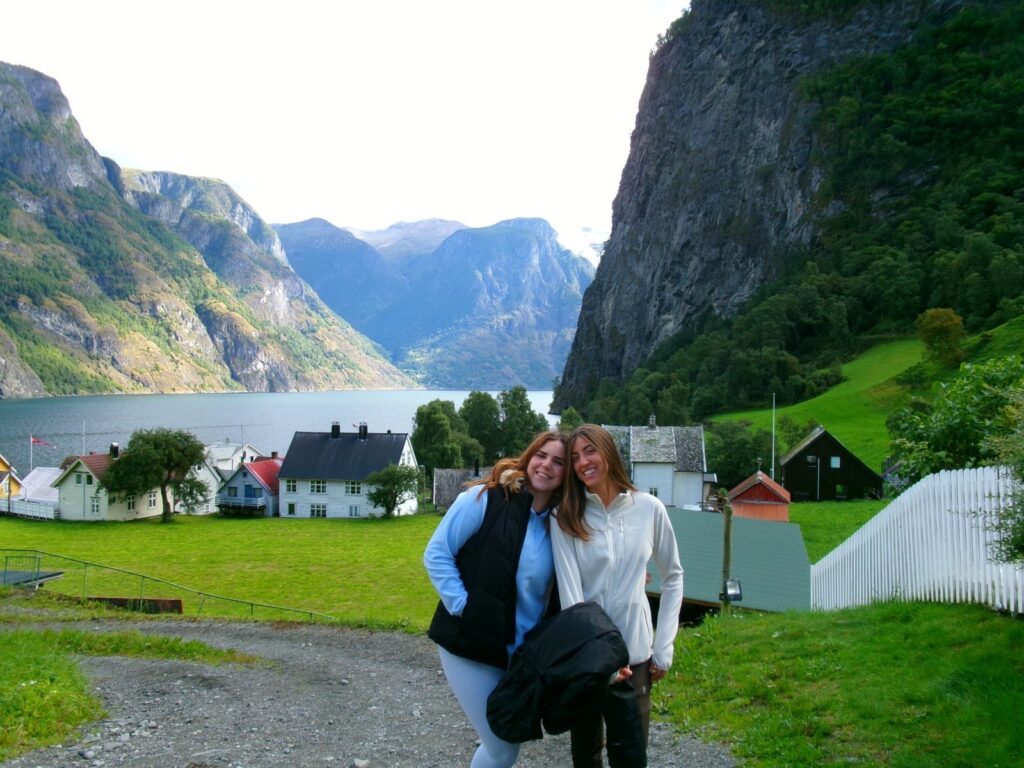 Dos amigas sonriendo junto a un pueblo de casas de madera a orillas de un fiordo noruego