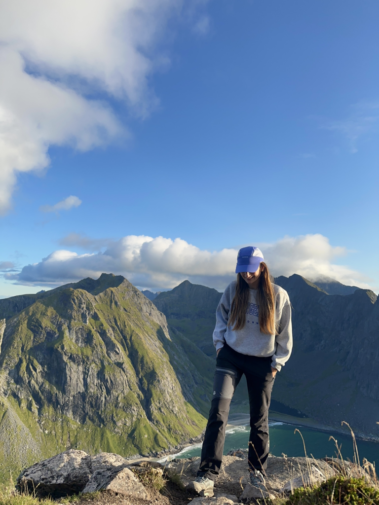 Mujer con gorra sonriendo en lo alto de una montaña de Lofoten, con picos verdes y una playa al fondo